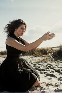 Side view of mid adult woman kneeling on sand at beach against sky