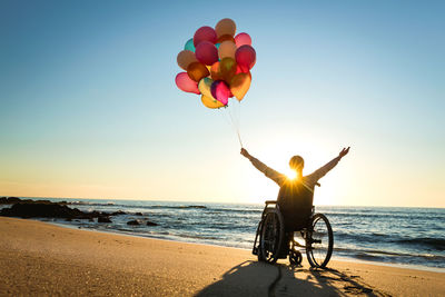 Silhouette mid adult woman with arms raised holding colorful balloons while sitting on wheelchair at beach during sunset