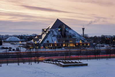 Snow covered illuminated buildings against sky during winter