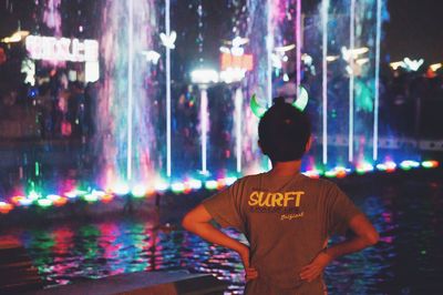 Rear view of boy viewing illuminated fountain at night in amusement park