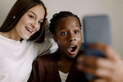 Shocked teenage girl looking at smart phone while sitting by smiling friend selfie at home