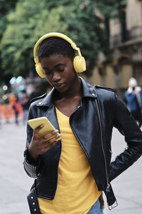 Young woman wearing headphones using smart phone while standing with hand on hip in city