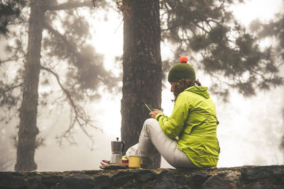Woman sitting on rock by tree trunk and coffee maker in forest during winter