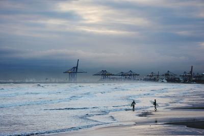 Surfers carrying surfboards on shore at beach against cloudy sky