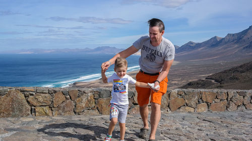 Full length of father and son walking against sea in sunny day