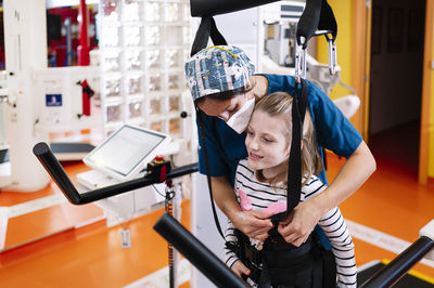 Professional medical specialist helping little girl with angelman syndrome and fixing straps while preparing for physiotherapy training on modern equipment in rehabilitation center