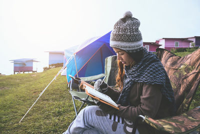 Side view of woman writing in diary while sitting on chair at campsite