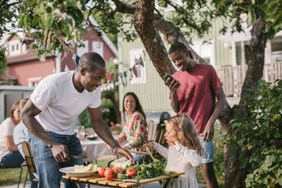 Boy photographing sister and father preparing food at table in backyard during garden party