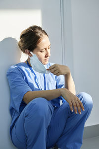 Nurse removing protective face mask squatting by wall in hospital