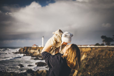 A woman is holding a baby near a lighthouse on the pacific coast