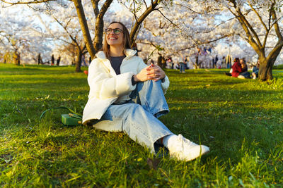 Woman in glasses, jeans and white jacket relaxing sitting under tree in cherry orchard in spring