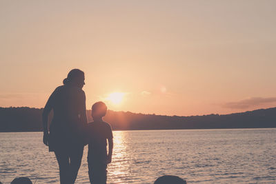 Rear view of silhouette woman with son standing in front of lake at sunset