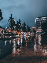 Wet illuminated street by buildings against sky during rainy season