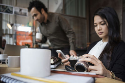 Businesswoman holding camera and phone with colleague in background at creative office