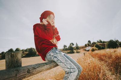 Woman sitting on railing in field against sky