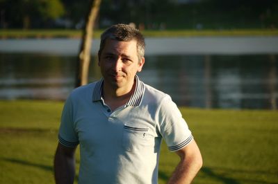 Portrait of man standing on field by lake during sunny day