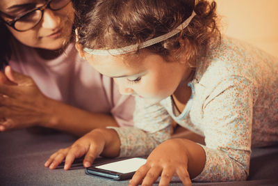 Small girl and her mother relaxing on the sofa and surfing the net on mobile phone.