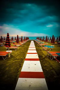 Empty boardwalk amidst lounge chairs at beach against cloudy sky