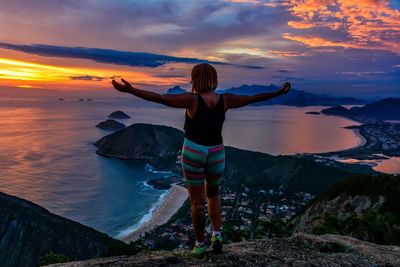 Rear view of woman with arms outstretched standing at beach during sunset