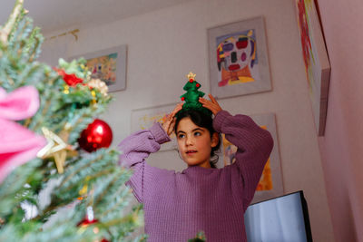 Standing behind decorated christmas tree girl adjusting the christmas headband on the head