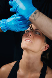 Cropped hands piercing woman eyebrow against black background