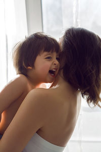 Mother in a white t-shirt holding her son in her arms standing at the window