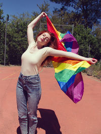 Young woman holding rainbow flag on basketball court against trees