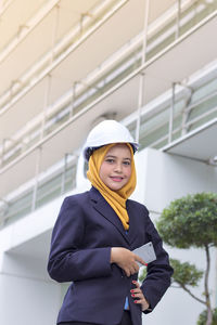 Low angle view of female architect holding smart phone while standing against office building