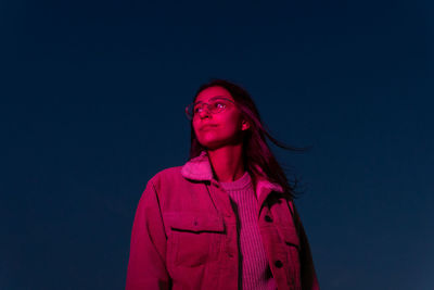 Attentive teen female with long brown hair in eyeglasses admiring dark night in pink illumination