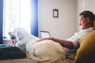 Senior man using laptop while leaning besides dog on bed at home