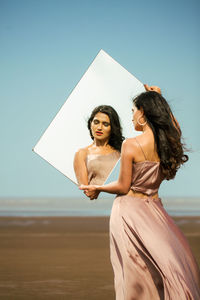 Woman holding mirror at beach against sky