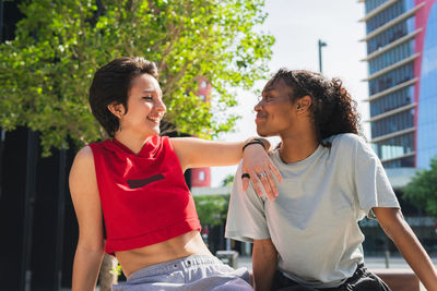 Cheerful multiracial young female friends in casual clothes looking at each other while sitting together on sunny street in city