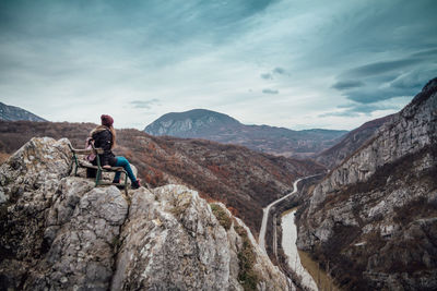 Woman sitting on rocks looking at mountains against sky