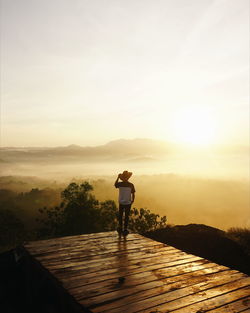 Rear view of man standing on boardwalk against sky during sunset