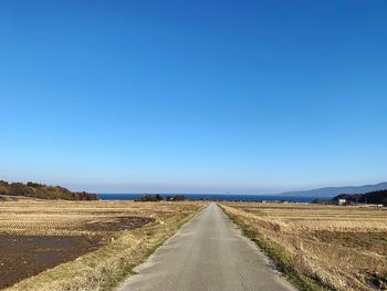 Empty road amidst field against clear blue sky