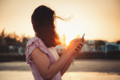 Side view of mid adult woman using smart phone while standing against sky during sunset