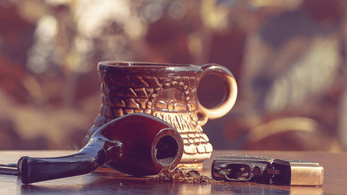 Close-up of cup with smoking pipe and lighter on table