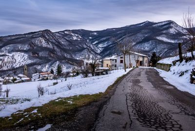 Snow covered road amidst buildings and mountains against sky