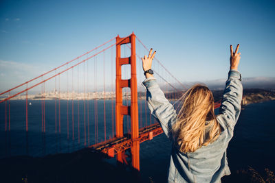 Rear view of woman gesturing by suspension bridge against sky