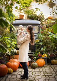 Woman playing with daughter while standing by pumpkins against trees