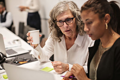 Senior businesswoman having coffee while discussing with colleague over laptop at office