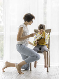 Mother cuts her son's hair by herself. little boy sits, covered with cloth,  holds pair of scissors.