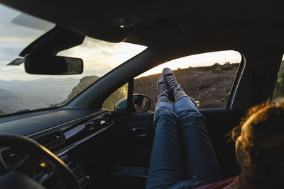 Young woman relaxing with feet up on car window at sunset