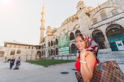 Portrait of smiling woman wearing headscarf standing against mosque in city