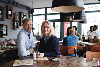 Portrait of smiling mature couple with menu at wooden table against people in restaurant