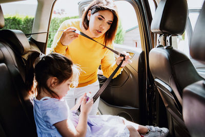 Young ethnic mother in stylish outfit fastening safety belt to little daughter using mobile phone on backseat of modern car on sunny day