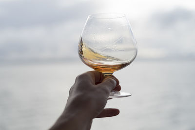 Cropped image of hand holding brandy snifter against sea