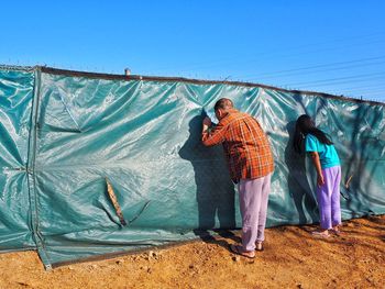 Man and girl peeking through fence against clear sky