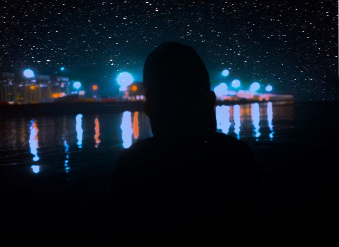 REAR VIEW OF SILHOUETTE MAN IN ILLUMINATED CITY AGAINST SKY