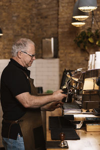 Side view of senior male barista in apron and eyeglasses putting portafilter in modern professional coffee machine while brewing espresso in cafe